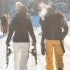 Im Vordergrund befinden zwei Menschen in Winterkleidung, von hinten fotografiert, die über einen verschneiten, sonnigen Waldweg laufen. Die Person links hält Schneeschuhe in den Händen. Im Hintergrund sind drei weitere Menschen zu sehen. 
